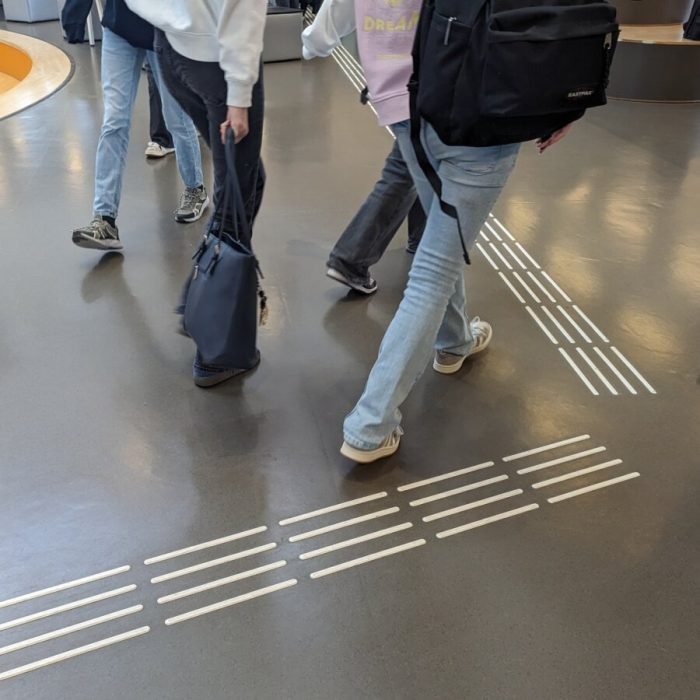 Kijkend naar de grond zie je een witte ribbels met een attentievlak waar de route van richting veranderd. Er lopen leerlingen overheen.